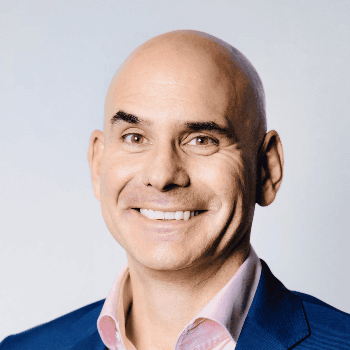 Holger Pfister headshot - A bald, tanned man smiling while wearing a pink shirt and a navy suit